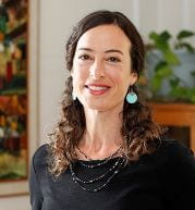 Laurie wears a black shirt and blue earrings. A plant and painting are visible behind her.