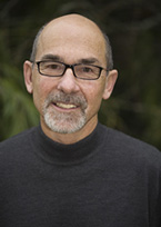 Don wears a black sweater and smiles, with foliage visible behind him.